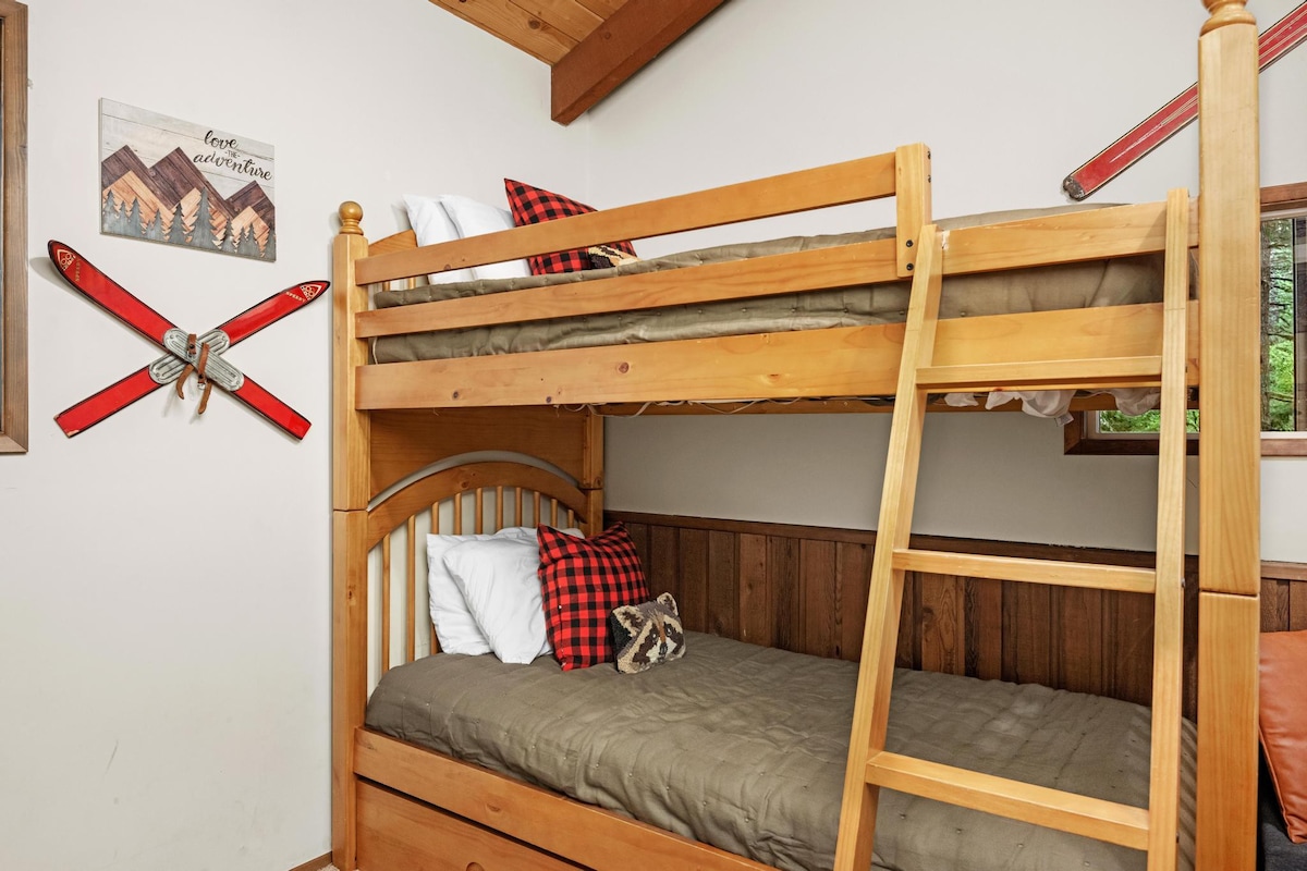 A detailed view of the wooden bunk bed in bedroom-1, featuring a sturdy ladder, plaid pillows, and a small window with a glimpse of greenery. Decorative red skis are mounted on the wall, enhancing the cabin theme.