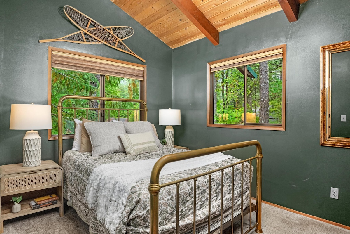 A serene bedroom with dark green walls and a brass-framed queen bed dressed in white and patterned bedding. Two large windows offer lush forest views, complemented by a rustic nightstand and decorative snowshoes.