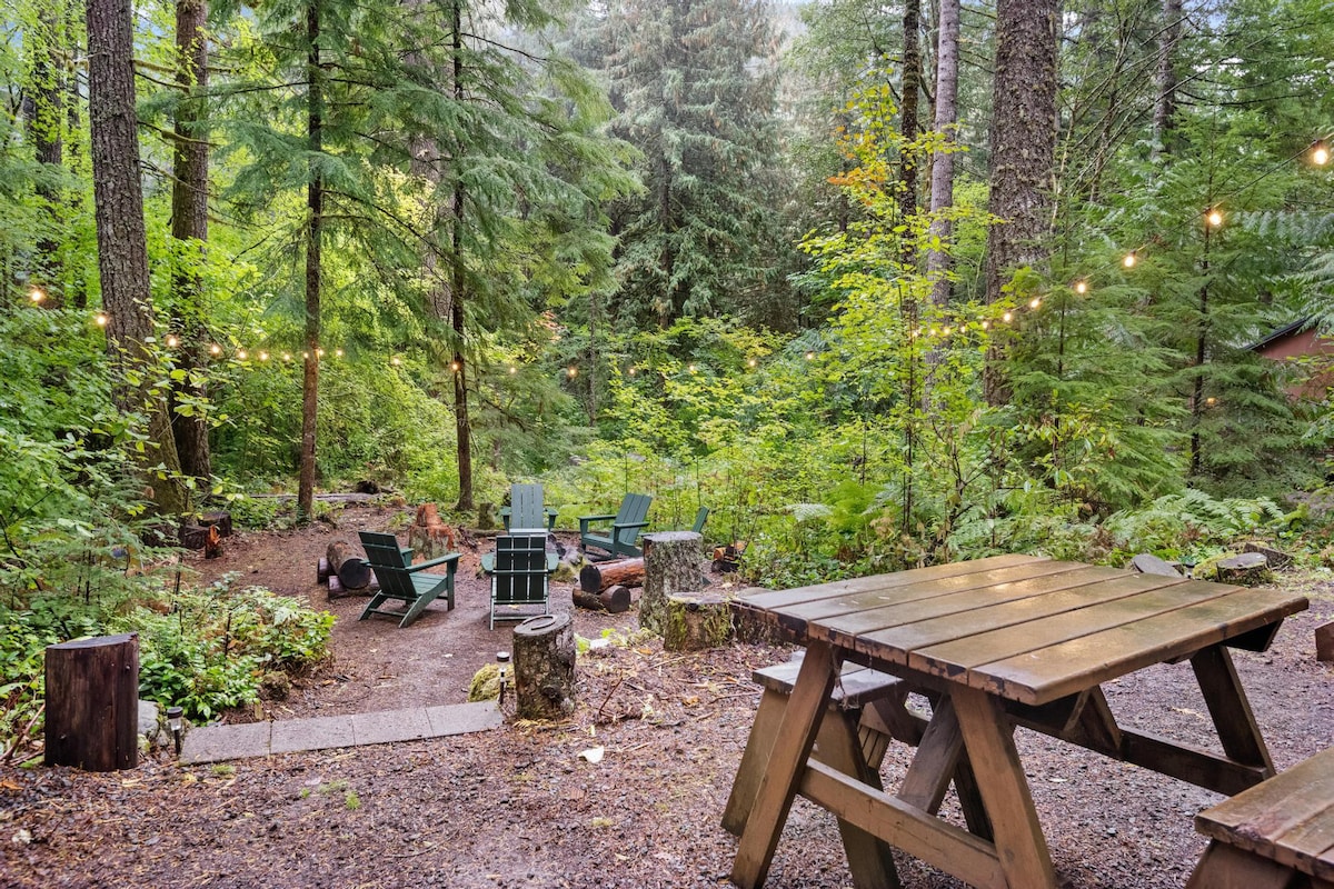 A rustic outdoor gathering space nestled in a lush forest, featuring a central fire pit surrounded by green Adirondack chairs and tree stump seating. String lights illuminate the area, with a wooden picnic table nearby.