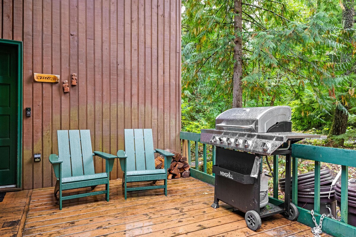 A wooden deck area next to the house, featuring two green Adirondack chairs and a large gas grill, perfect for outdoor cooking. The surrounding area is lush with trees, and stacked firewood is visible.