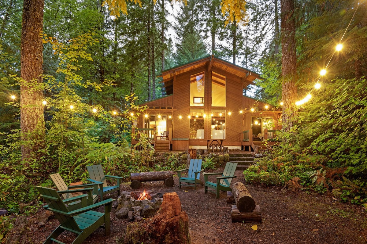 The charming wooden exterior of the house, illuminated by string lights, is set against a vibrant forest backdrop. The outdoor fire pit area with Adirondack chairs and a picnic table is visible in the foreground.