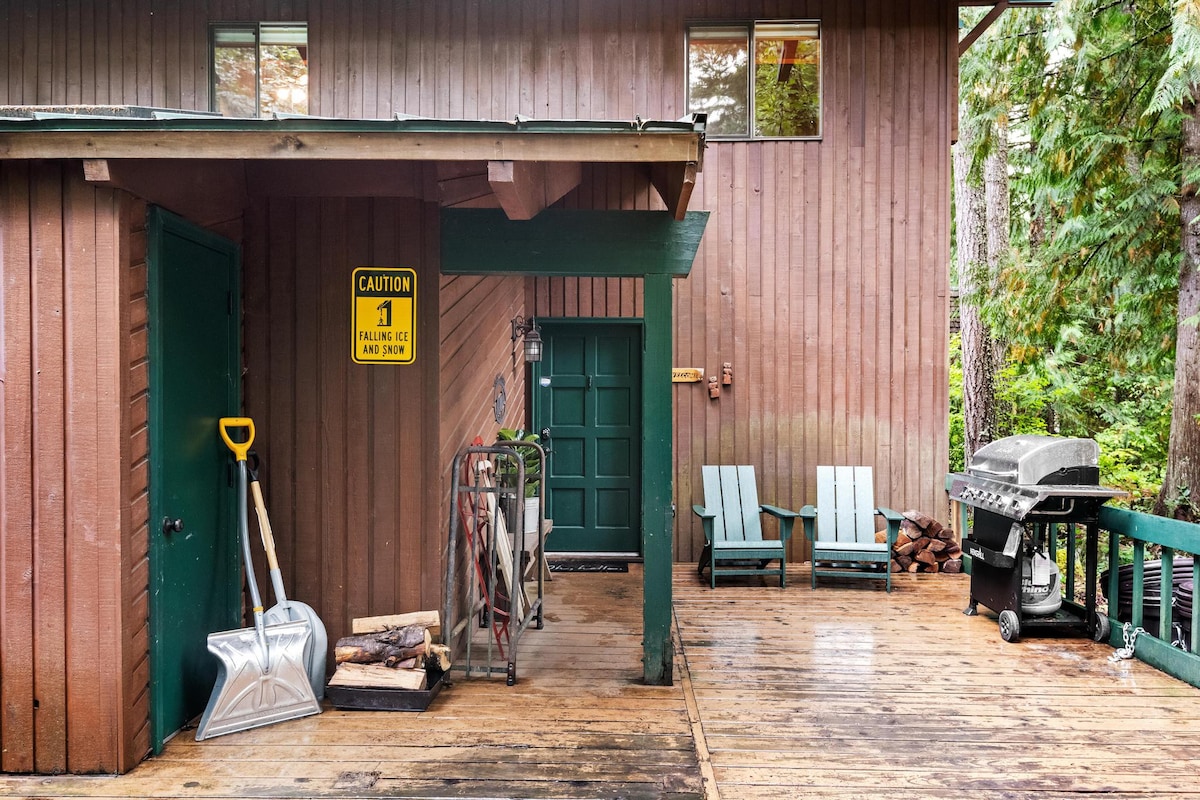 A wooden deck area next to the cabin's side entrance, featuring a gas grill, two light blue Adirondack chairs, and a "Caution Falling Ice and Snow" sign. Shovels and firewood are stored near a green door.