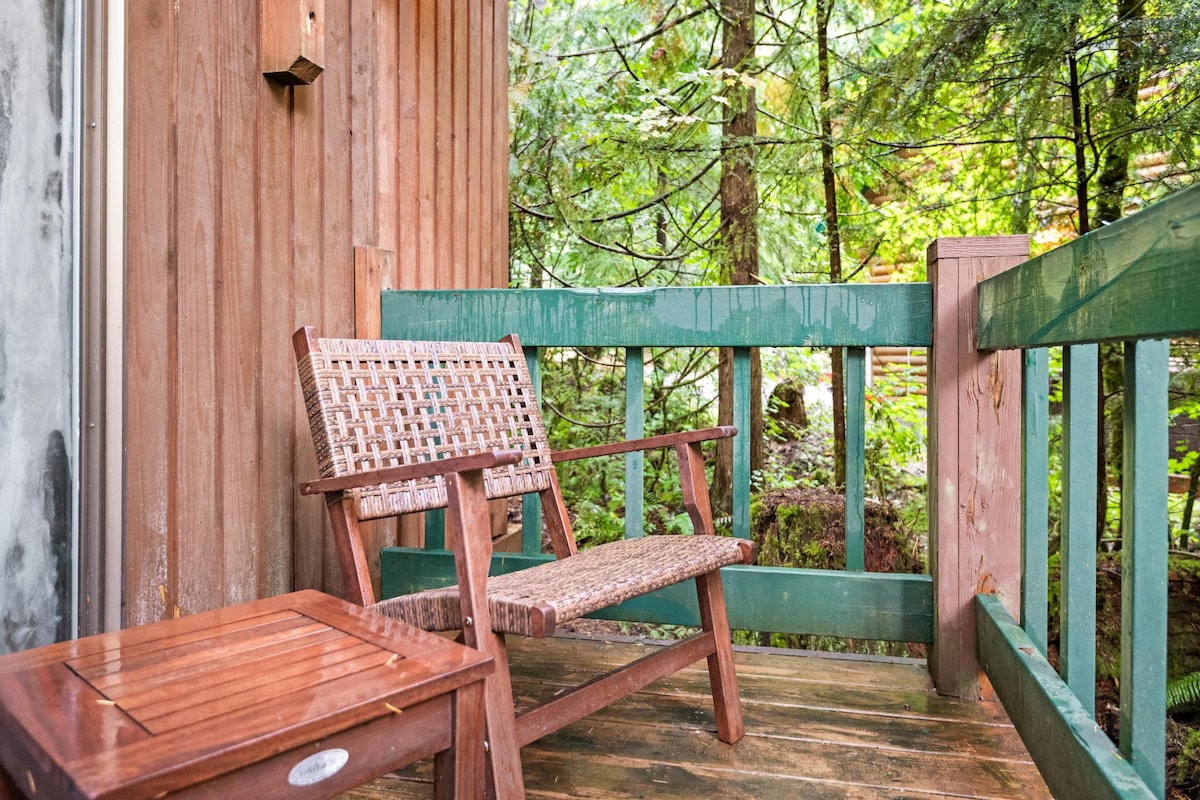 A cozy wooden balcony with a woven chair and a small side table, offering a serene view of the surrounding dense green forest. A green railing encloses the space.
