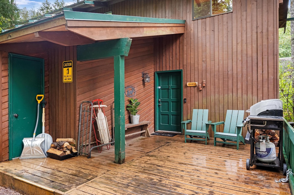 A spacious wooden deck extends from the cabin's side, equipped with a gas grill, two light blue Adirondack chairs, and a bench. A green door serves as an entrance, with firewood and snow equipment nearby.