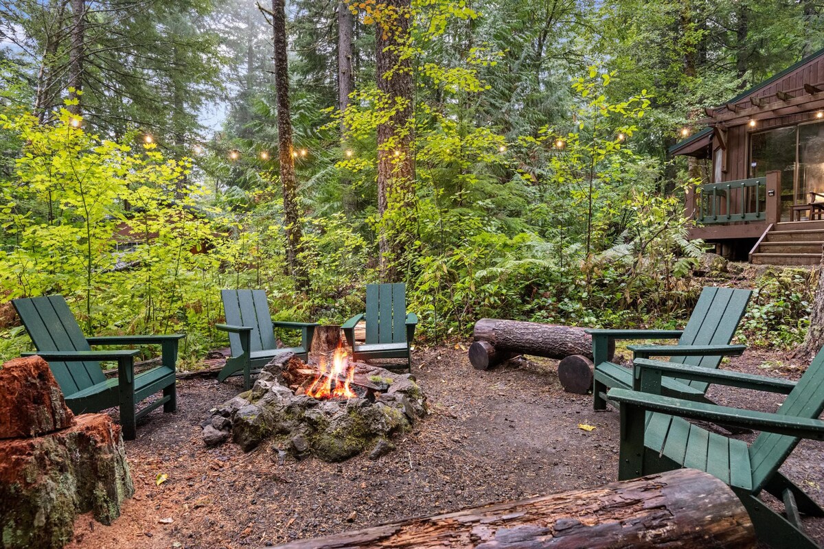 A rustic fire pit area with a crackling fire, surrounded by four dark green Adirondack chairs and log benches. The setting is nestled in a lush forest with tall trees.