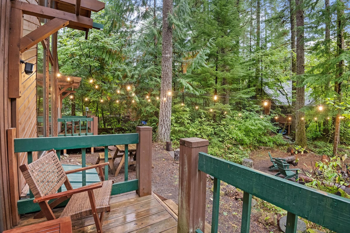 Elevated view from a wooden deck, showing a fire pit area below with Adirondack chairs and log seating, all illuminated by string lights strung through the surrounding forest.