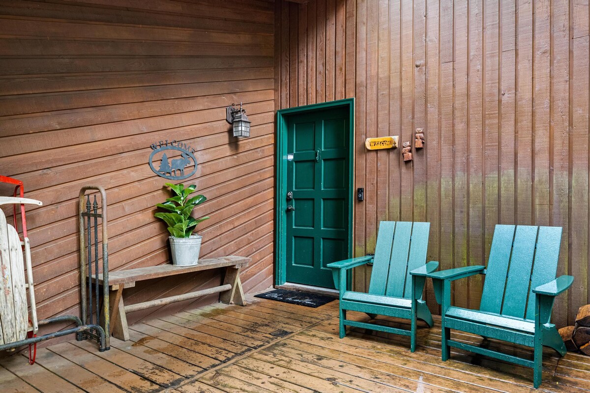 A welcoming wooden porch area leading to a dark green entrance door, flanked by two light blue Adirondack chairs and a wooden bench with a potted plant. A "Welcome" sign adorns the wooden siding.
