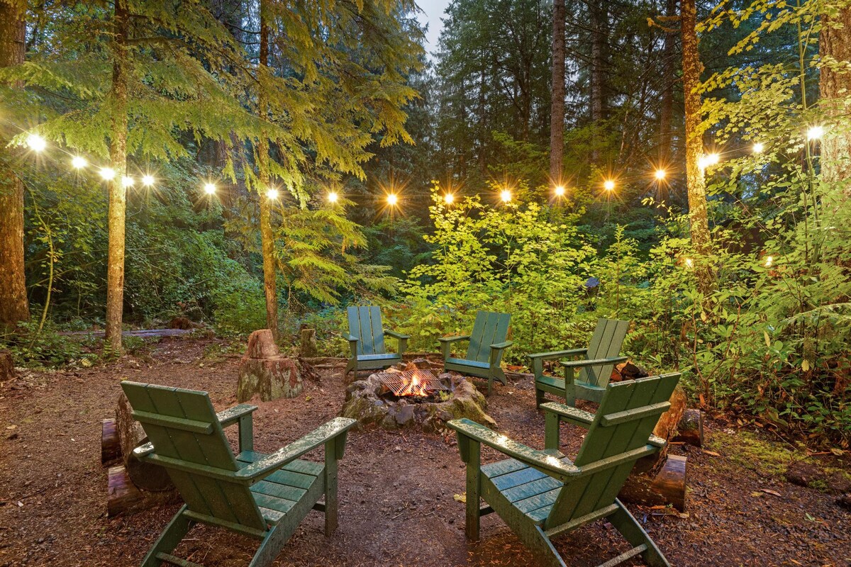 Outdoor fire pit area nestled in a lush forest, featuring six green Adirondack chairs arranged around a central fire pit, illuminated by string lights strung between tall trees.
