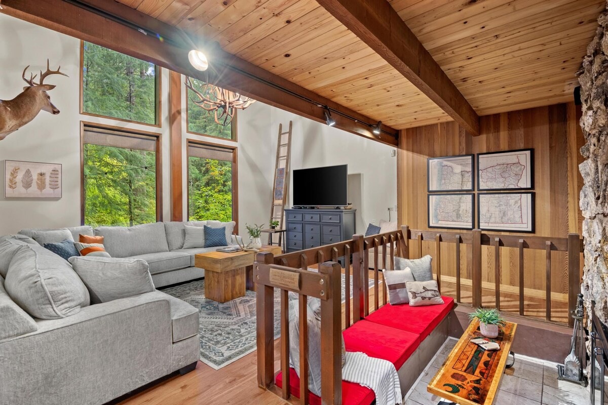 A wide shot of the living room featuring a large gray sectional sofa, a rustic wooden coffee table, and floor-to-ceiling windows with forest views, connected to a lower level by a wooden railing.