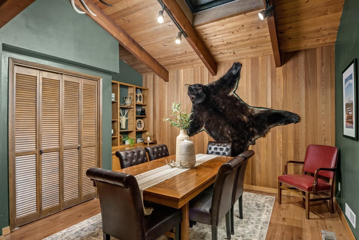 A rustic dining room with a wooden table and six brown leather chairs, featuring wood-paneled walls, a vaulted ceiling, and a large black bear rug mounted on the wall.