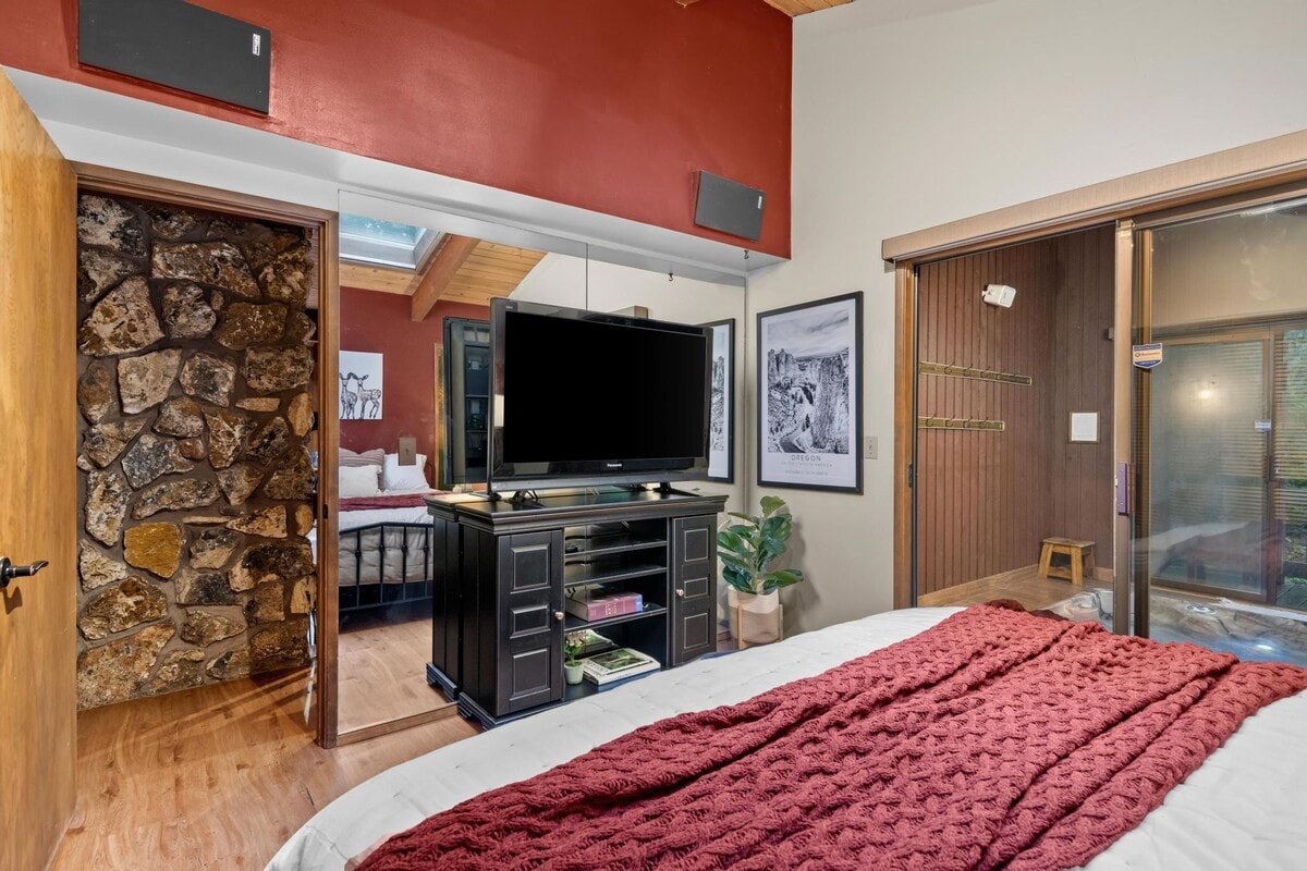 View of the bedroom showing a flat-screen TV on a dark media console at the foot of the bed, with a mirrored closet door reflecting the room, and a stone-accented wall visible through an open doorway.