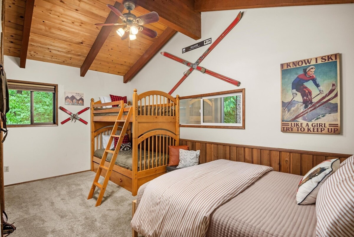 A spacious bedroom combining a wooden bunk bed and a queen bed, both with cozy bedding. The room boasts a vaulted wooden ceiling, a ceiling fan, and ski-themed decor, including a vintage poster and mounted skis.