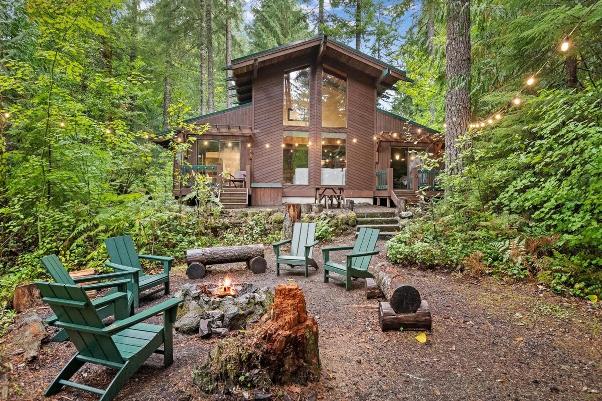 A rustic wooden cabin nestled in a lush green forest, featuring a prominent fire pit area with green Adirondack chairs and log seating in the foreground. String lights illuminate the trees and the cabin's large windows.