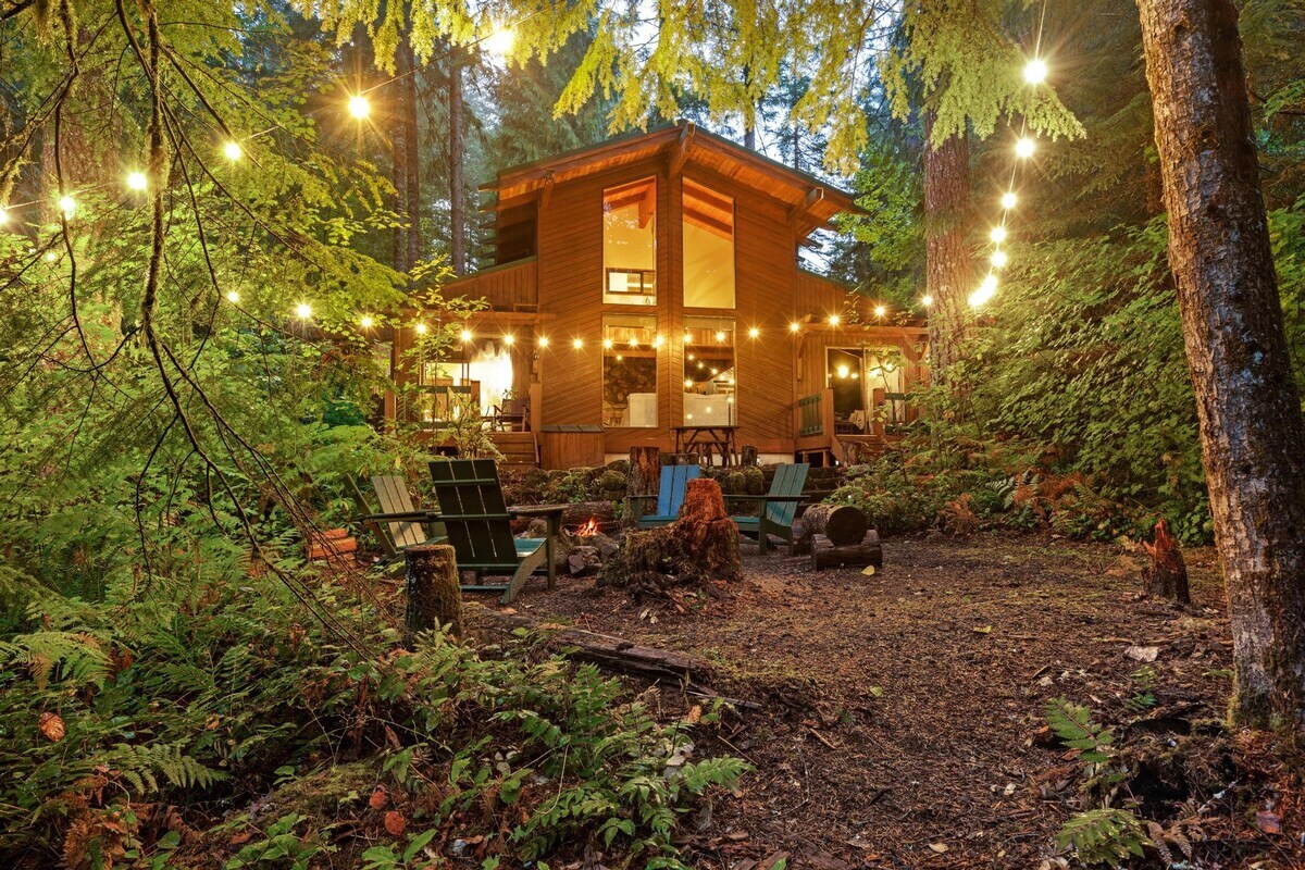 The charming wooden cabin is illuminated at dusk by warm interior lights and festive string lights draped through the surrounding trees. A fire pit area with seating is visible in the foreground, set within the lush forest.